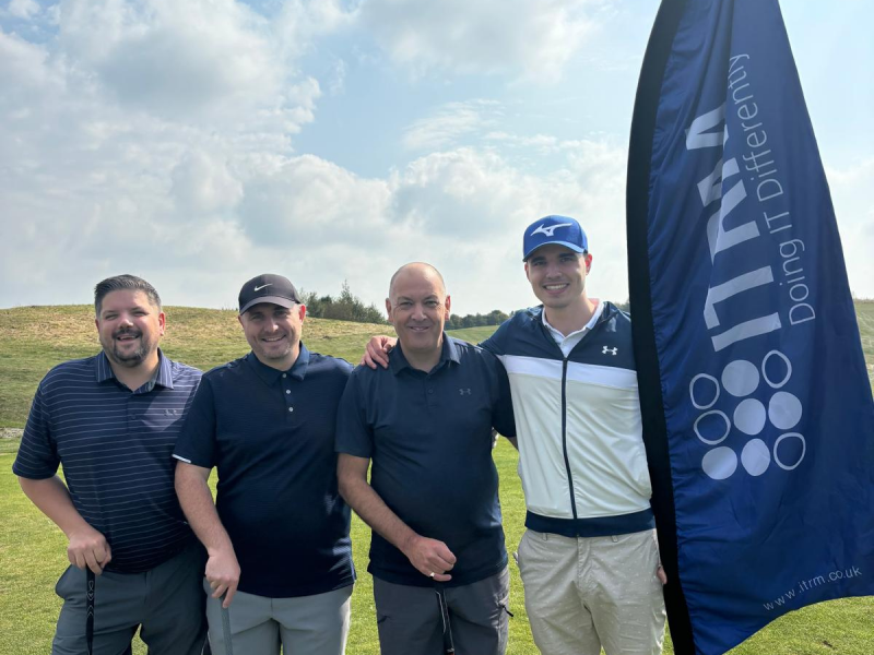 Four ITRM staff standing together on a golf course, smiling and holding golf clubs. They are dressed in casual golf attire with a blue ITRM flag in the background displaying the slogan 'Doing IT Differently.' The sky is partly cloudy with patches of blue,